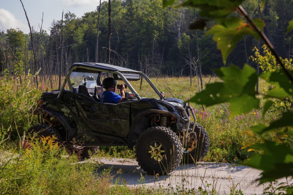 A two-up side-by-side off road vehicle is stopped on a gravel trail beside a wetland. The driver is taking a photo of something in the distance with a professional looking camera.