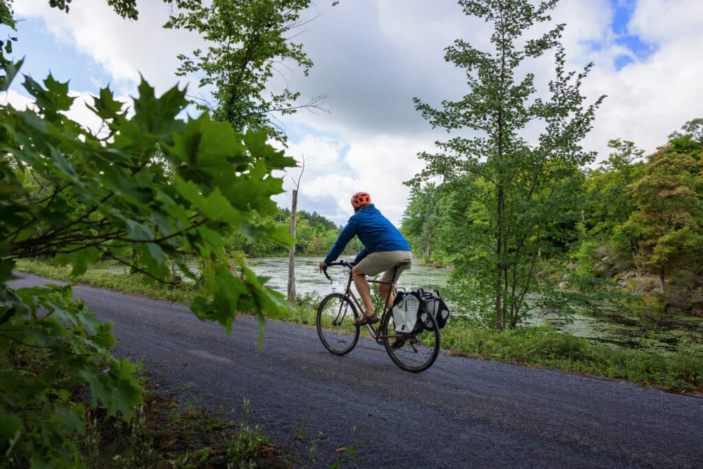A touring cyclist travels on a gravel travel past a wetland, surrounded by lush green forest.