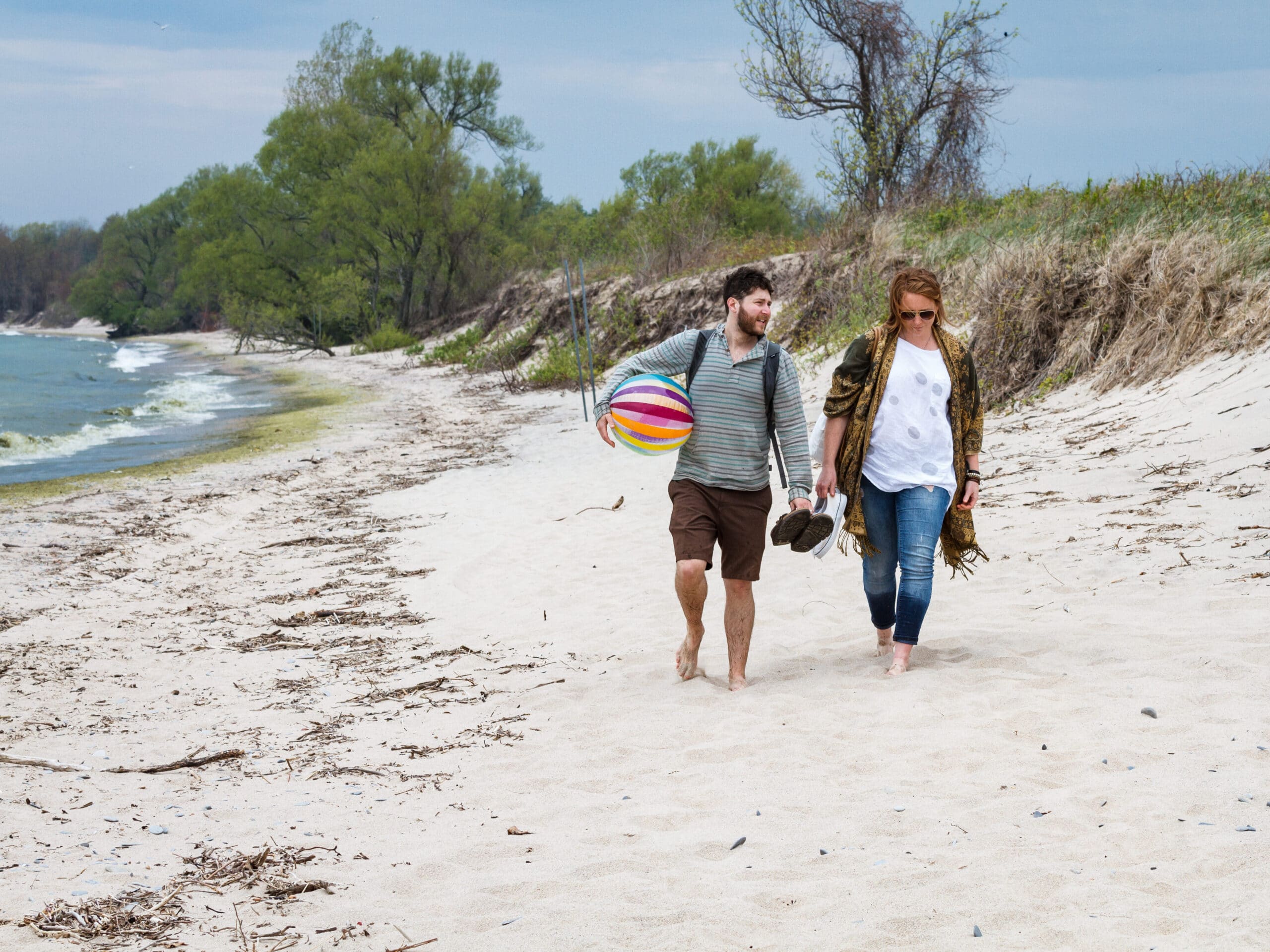 A bearded man and a woman, both decked out in sunglasses carry their shoes and a beach ball as they walk along a white sandy beach in their bare feet.