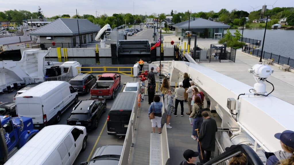 A few vans, trucks and cars wait for the Wolfe Island Ferry to dock at Marysville with the terminal and village in the background. A group of pedestrians watch the docking procedure from the observation deck.