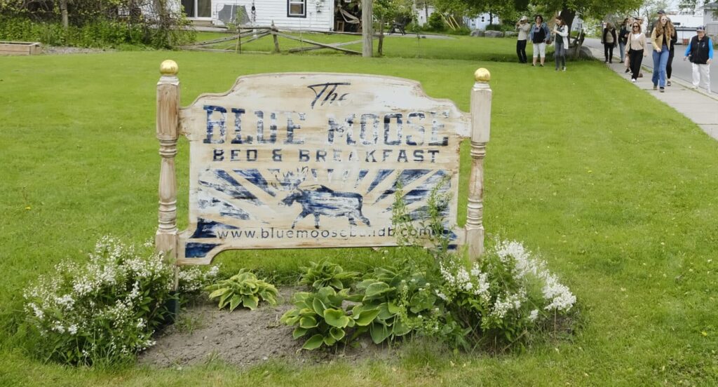 Sign reading Blue Moose Bed and Breakfast with a group of people walking down a street behind it.
