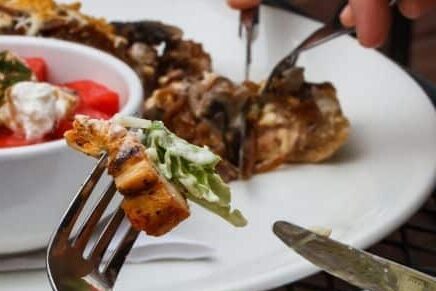 A fork with a portion of a gourmet meal, where additional food is being cut on a plate in the background.