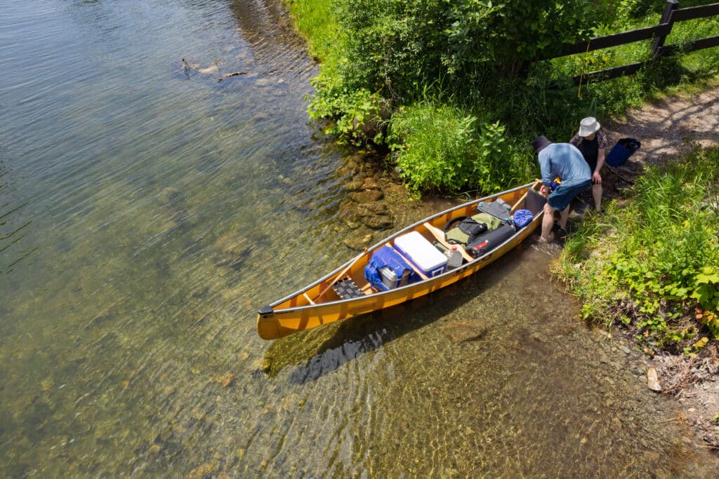 Two people are launching a loaded yellow canoe into a clear lake on warm, sunny day.